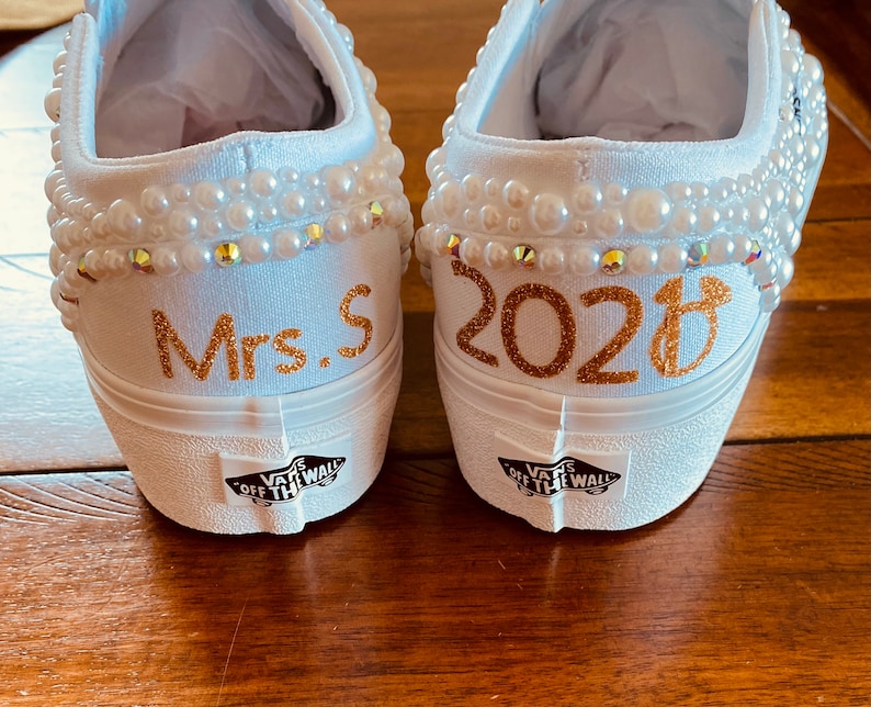 Puede incluir: Un par de zapatos de lona blancos con adornos de perlas. Un zapato dice "Mrs. S" y el otro dice "2023".