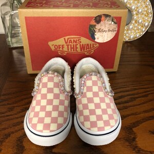 Puede incluir: Un par de zapatos Vans slip-on a cuadros rosa y blanco con adornos de perlas alrededor de los bordes. Los zapatos son nuevos y est&aacute;n en su caja original. La caja es marr&oacute;n con el logotipo de Vans y las palabras "Off The Wall" impresas en ella.