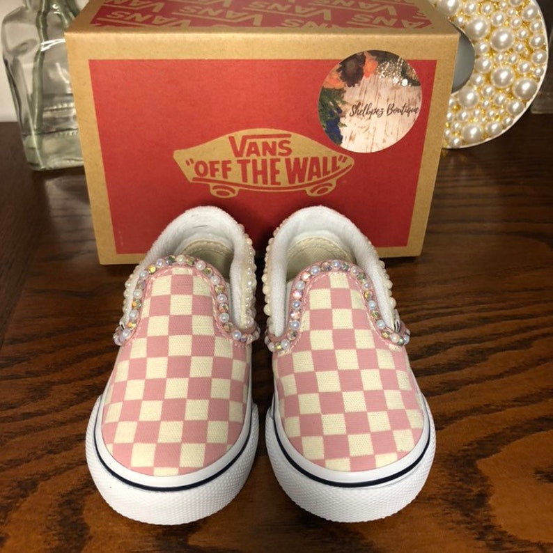 Puede incluir: Un par de zapatos Vans slip-on a cuadros rosa y blanco con adornos de perlas alrededor de los bordes. Los zapatos son nuevos y est&aacute;n en su caja original. La caja es marr&oacute;n con el logotipo de Vans y las palabras "Off The Wall" impresas en ella.