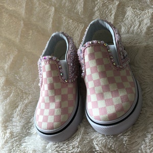 Puede incluir: Un par de zapatos Vans de slip-on a cuadros rosa y blanco con un dise&ntilde;o adornado. Los zapatos tienen una suela blanca y un borde negro.