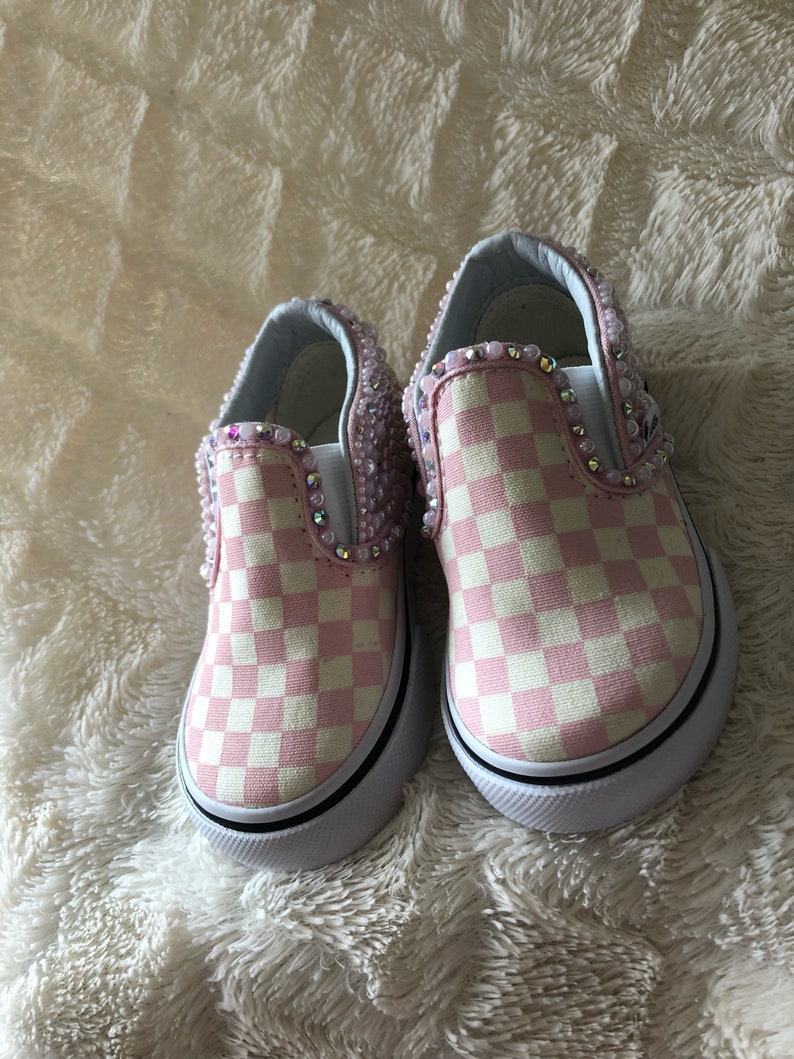 Puede incluir: Un par de zapatos Vans de slip-on a cuadros rosa y blanco con un dise&ntilde;o adornado. Los zapatos tienen una suela blanca y un borde negro.