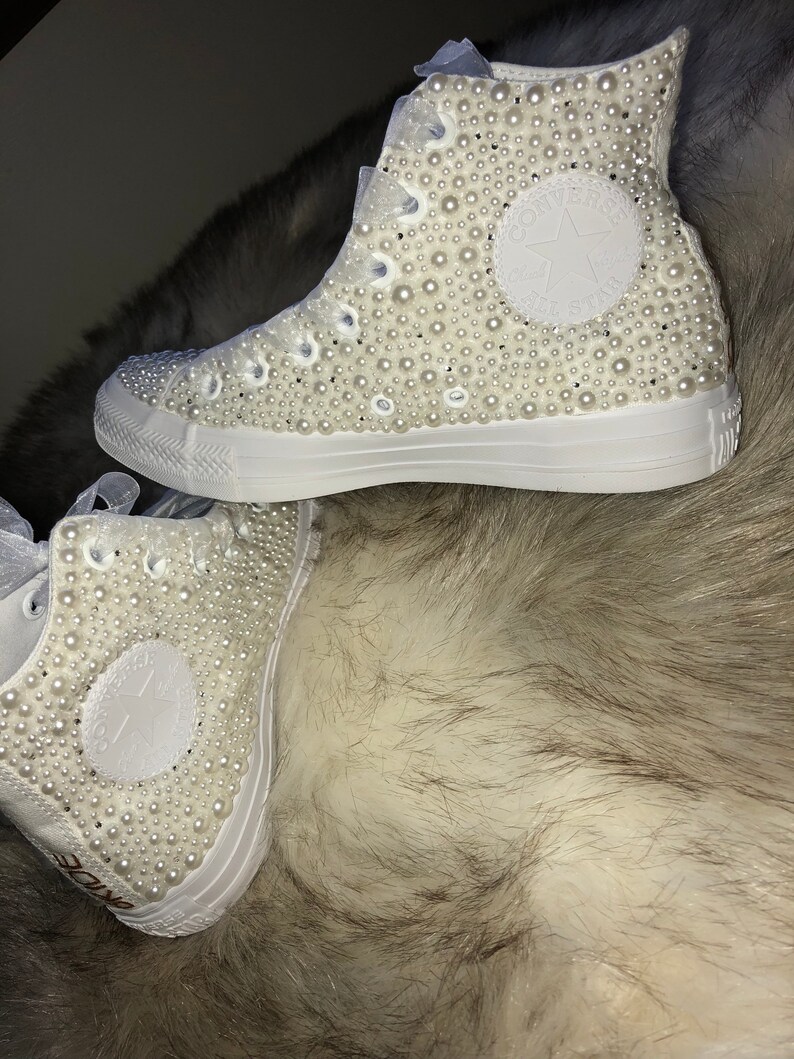 May include: White high-top sneakers covered in white pearl embellishments. The sneakers have a white star logo on the side.