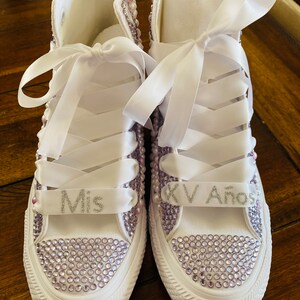 May include: White high-top sneakers with lavender rhinestones and white satin ribbon laces. The left shoe has "Mis" written on the ribbon and the right shoe has "XV A&ntilde;os" written on the ribbon.