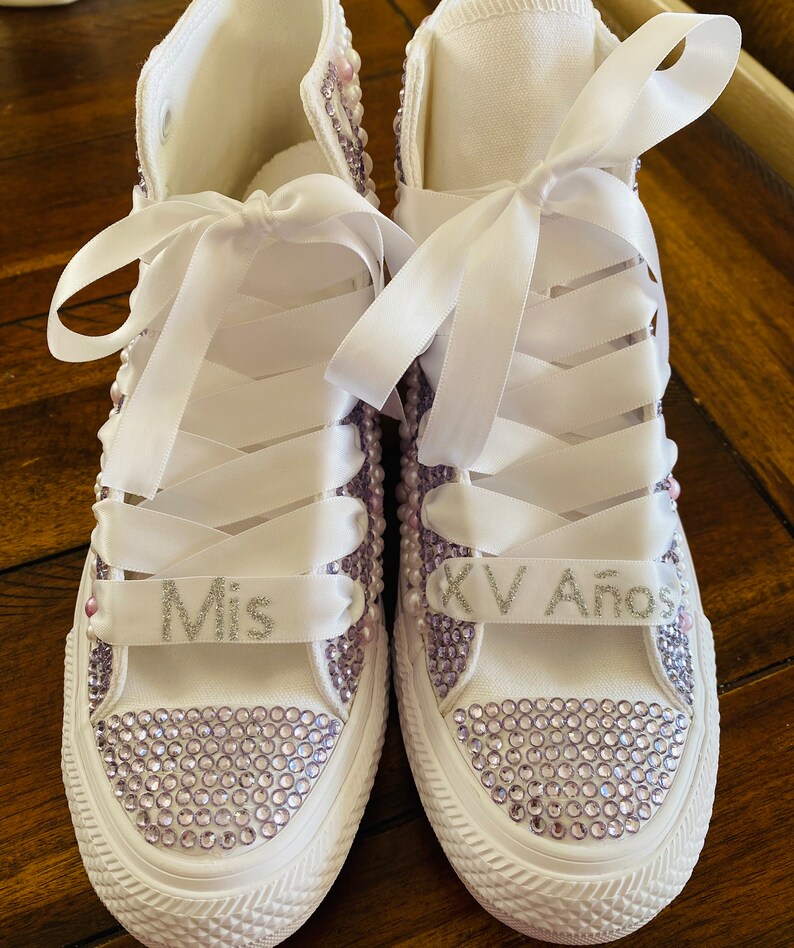 May include: White high-top sneakers with lavender rhinestones and white satin ribbon laces. The left shoe has "Mis" written on the ribbon and the right shoe has "XV A&ntilde;os" written on the ribbon.
