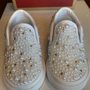 Puede incluir: Un par de zapatillas blancas de lona con un adorno de perlas y pedrer&iacute;a. Las zapatillas tienen una suela de goma blanca y una parte superior de lona blanca.