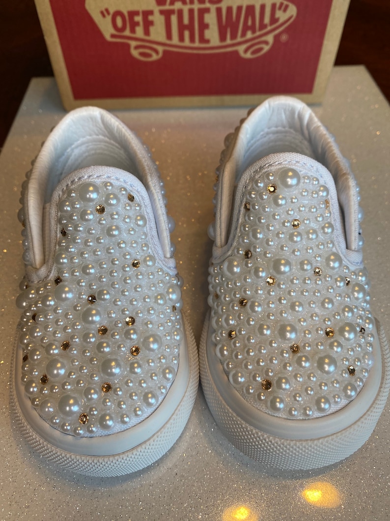 Puede incluir: Un par de zapatillas blancas de lona con un adorno de perlas y pedrer&iacute;a. Las zapatillas tienen una suela de goma blanca y una parte superior de lona blanca.