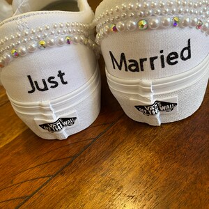 Puede incluir: Un par de zapatos de lona blancos con adornos de perlas. Un zapato dice "Just" y el otro dice "Married".