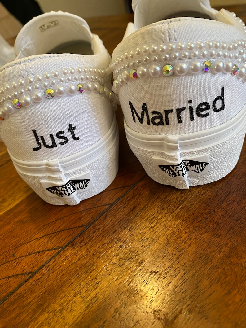 Puede incluir: Un par de zapatos de lona blancos con adornos de perlas. Un zapato dice "Just" y el otro dice "Married".
