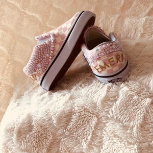 Puede incluir: Un par de zapatos Vans slip-on a cuadros rosa y blanco con adornos de perlas y el nombre "EMERY" escrito en purpurina dorada en la parte posterior de un zapato.