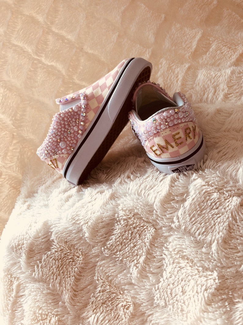 Puede incluir: Un par de zapatos Vans slip-on a cuadros rosa y blanco con adornos de perlas y el nombre "EMERY" escrito en purpurina dorada en la parte posterior de un zapato.