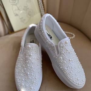 Puede incluir: Un par de zapatillas blancas de tela sin cordones cubiertas de perlas blancas. Las zapatillas est&aacute;n sobre una superficie de color marr&oacute;n claro.