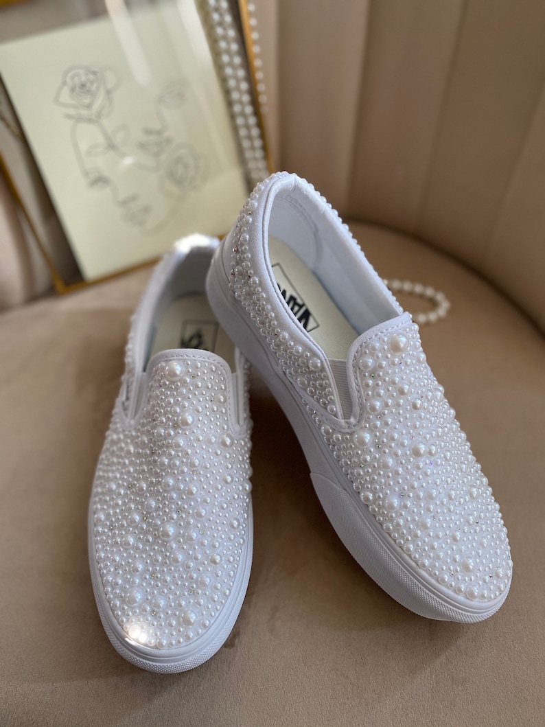 Puede incluir: Un par de zapatillas blancas de tela sin cordones cubiertas de perlas blancas. Las zapatillas est&aacute;n sobre una superficie de color marr&oacute;n claro.