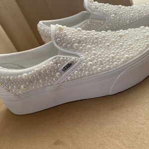 Puede incluir: Zapatillas Vans blancas de slip-on con suela de plataforma, cubiertas de adornos de perlas blancas. Las zapatillas tienen el logotipo de Vans en el lateral.