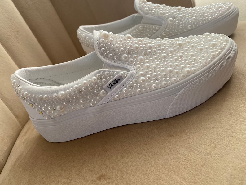 Puede incluir: Zapatillas Vans blancas de slip-on con suela de plataforma, cubiertas de adornos de perlas blancas. Las zapatillas tienen el logotipo de Vans en el lateral.