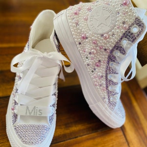 May include: A pair of white high-top sneakers with a white satin ribbon and pearl and crystal embellishments. The left shoe has the word "Mis" written on the ribbon.