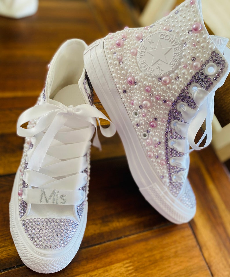 May include: A pair of white high-top sneakers with a white satin ribbon and pearl and crystal embellishments. The left shoe has the word "Mis" written on the ribbon.