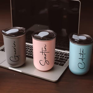 Puede incluir: Tres tazas de viaje de acero inoxidable con nombres personalizados: "Gwerin", "Sartin" y "Odusta". Las tazas están sobre un teclado de portátil con un esquema de color gris, negro, rosa y azul.