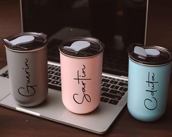 May include: Three stainless steel travel mugs with personalized names: "Gwerin", "Sartin", and "Odusta". The mugs are on a laptop keyboard with a gray, black, pink, and blue color scheme.