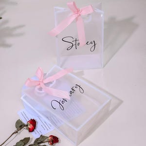 Puede incluir: Dos bolsas de regalo de pl&aacute;stico transparente con lazos de cinta rosa. Las bolsas est&aacute;n decoradas con caligraf&iacute;a negra. Una bolsa dice "Stacey" y la otra dice "Enero".