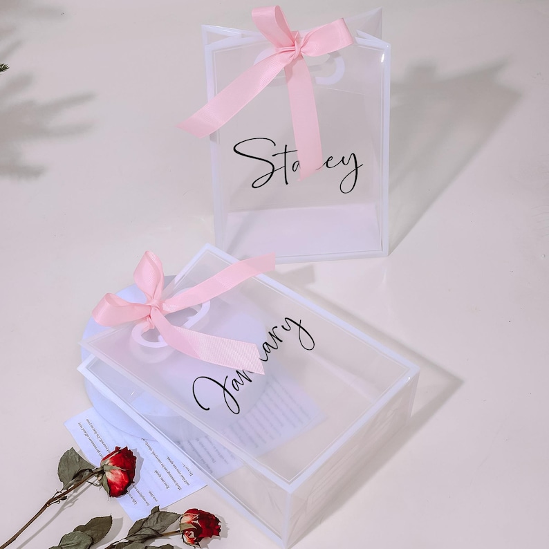 Puede incluir: Dos bolsas de regalo de pl&aacute;stico transparente con lazos de cinta rosa. Las bolsas est&aacute;n decoradas con caligraf&iacute;a negra. Una bolsa dice "Stacey" y la otra dice "Enero".