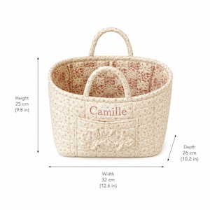 May include: A quilted fabric basket with a floral pattern, featuring two handles, a front pocket with a ruffled trim, and the name Camille embroidered in pink. The basket measures 9.8 inches in height, 12.6 inches in width, and 10.2 inches in depth.