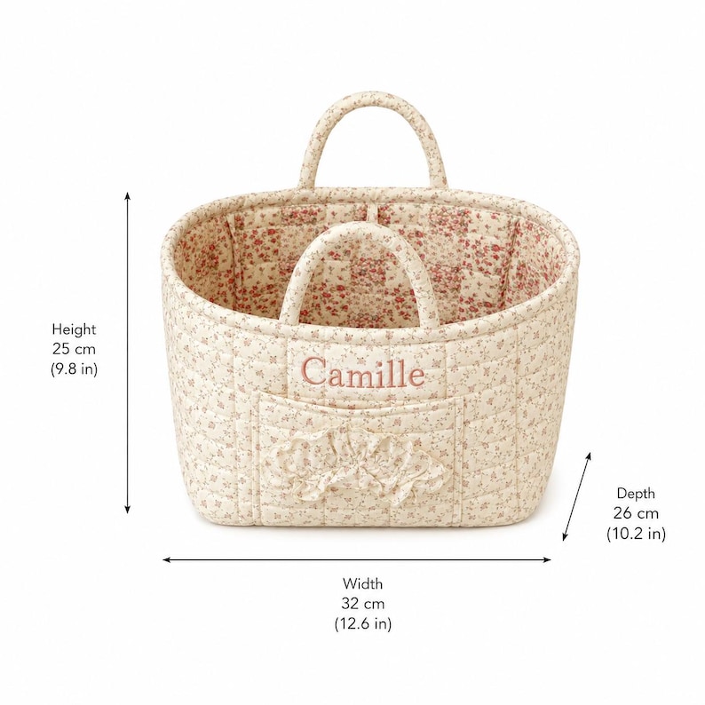 May include: A quilted fabric basket with a floral pattern, featuring two handles, a front pocket with a ruffled trim, and the name Camille embroidered in pink. The basket measures 9.8 inches in height, 12.6 inches in width, and 10.2 inches in depth.