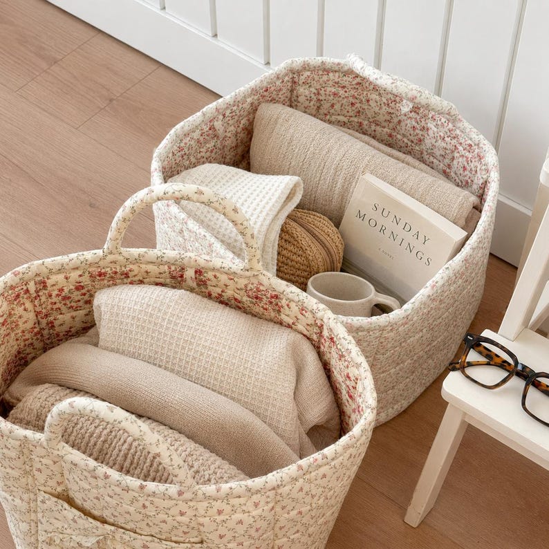 May include: Two quilted fabric baskets with a cream and red floral pattern, each featuring two handles. The baskets contain folded beige textiles, a small tan pouch, a white mug, and a book titled Sunday Mornings. Tortoiseshell glasses sit nearby.