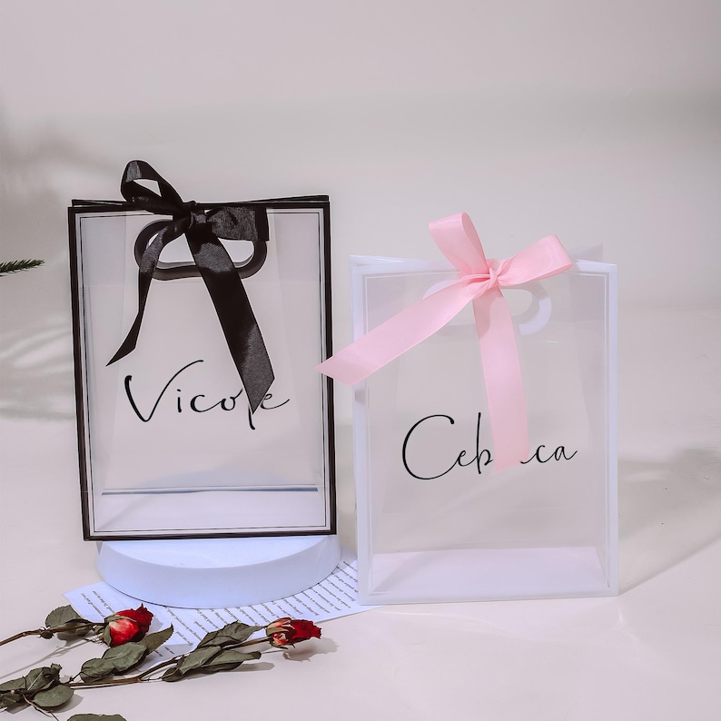 Puede incluir: Dos bolsas de regalo de pl&aacute;stico transparente con lazos de cinta negra y rosa. La bolsa de la izquierda tiene una cinta negra y el texto "Vicole" impreso en la parte delantera. La bolsa de la derecha tiene una cinta rosa y el texto "Celca" impreso en la parte delantera.