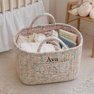 May include: A quilted fabric storage basket featuring a colorful floral pattern, two handles, and a front pocket with a ruffled trim. The name Ava is embroidered in dark brown thread on the front. Inside are folded cream garments and two books.