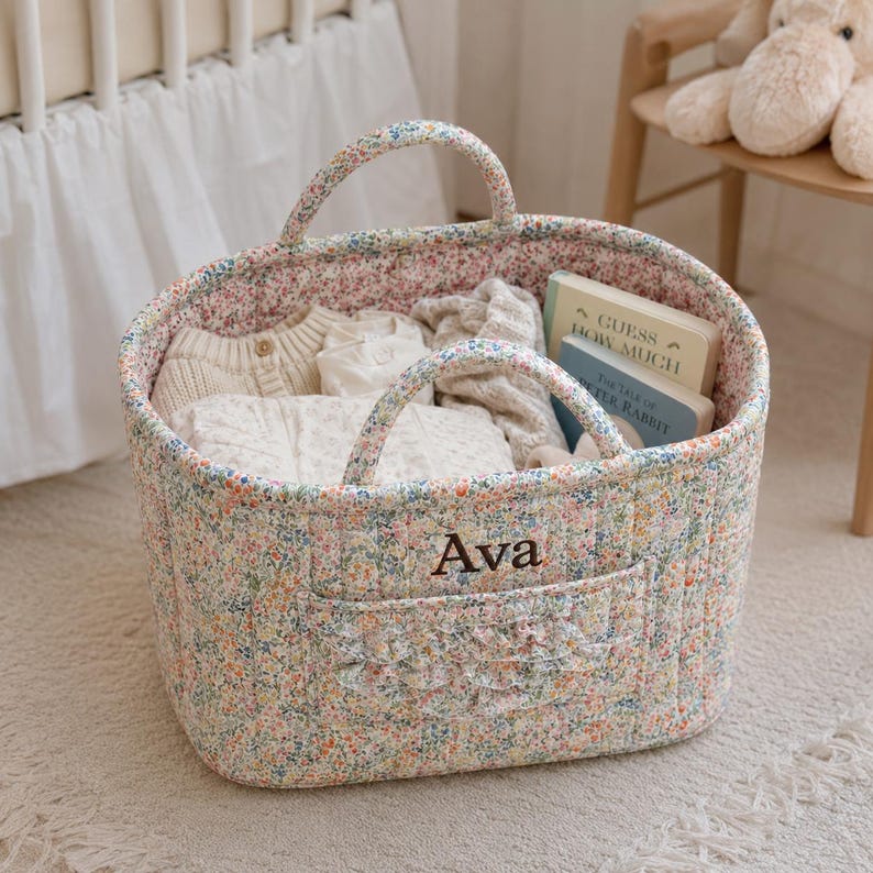 May include: A quilted fabric storage basket featuring a colorful floral pattern, two handles, and a front pocket with a ruffled trim. The name Ava is embroidered in dark brown thread on the front. Inside are folded cream garments and two books.