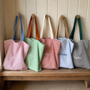 Puede incluir: Cinco bolsas de tela con rayas verticales en rosa, verde claro, rojo, azul claro y negro. Cada bolsa tiene un nombre bordado en letra cursiva blanca o azul: Laura, Mia, Ava, Emma y Sophie. Las bolsas tienen asas de colores a juego.