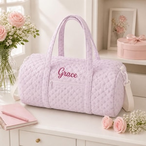 Könnte beinhalten: Eine hellviolette, gesteppte Reisetasche mit Rautenmuster und dem in Dunkelrosa gestickten Namen Grace auf der Vorderseite. Die Tasche verfügt über zwei Tragegriffe, eine Reißverschlusstasche auf der Vorderseite und weiße Befestigungselemente.