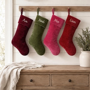 Puede incluir: Cuatro calcetines navideños de terciopelo colgados en fila, cada uno con un puño doblado bordado con un nombre. Los calcetines son de color burdeos oscuro, verde oliva, rosa brillante y rojo vibrante, con los nombres Ava, Noah, Mia y Liam.