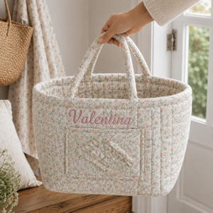 May include: A quilted fabric basket featuring a small-scale floral pattern in soft pink, green, and blue tones. The basket has two top handles and a rectangular front pocket. The name Valentina is embroidered in pink script on the front.