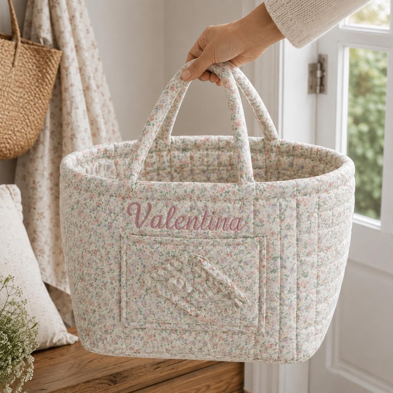 May include: A quilted fabric basket featuring a small-scale floral pattern in soft pink, green, and blue tones. The basket has two top handles and a rectangular front pocket. The name Valentina is embroidered in pink script on the front.
