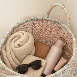 May include: A quilted fabric basket featuring a small-scale floral print in shades of pink, blue, and green. Inside the basket are a cream-colored sherpa fleece blanket, a tan wallet, a matte beige water bottle, sunglasses, and a small tube.