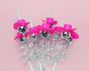 May include: A collection of cocktail stirrers with a disco ball and hot pink cowboy hat design. The stirrers have clear plastic stems and a decorative, swirled base. The background is a solid, light pink.