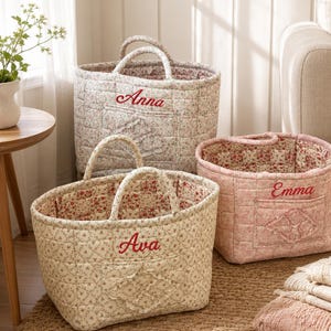 Puede incluir: Tres cestas de almacenamiento de tela acolchada con dos asas cada una. Las cestas presentan estampados florales en tonos crema y rosa. Cada cesta tiene un bolsillo frontal con un detalle de rombo cosido y está bordada con un nombre: Anna, Ava o Emma.