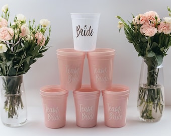 May include: Six pink plastic cups with the text "Team Bride" printed on them. A white plastic cup with the text "Bride" is on top of the cups.
