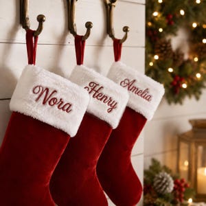 Peut inclure: Trois chaussettes de Noël en velours rouge suspendues à des crochets en métal couleur bronze. Chaque chaussette présente un revers en fausse fourrure blanche brodé d'un prénom en écriture rouge : Nora, Henry et Amelia.