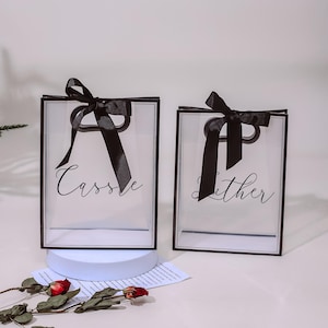 Puede incluir: Dos bolsas de regalo de pl&aacute;stico transparente con lazos de cinta negra y asas negras. Las bolsas tienen bordes negros y los nombres "Cassie" y "Lither" impresos en ellas.