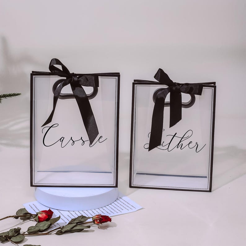 Puede incluir: Dos bolsas de regalo de pl&aacute;stico transparente con lazos de cinta negra y asas negras. Las bolsas tienen bordes negros y los nombres "Cassie" y "Lither" impresos en ellas.