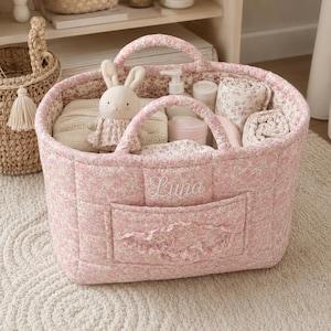 May include: A quilted pink fabric basket with a floral pattern and two handles. The front features a pocket with a ruffled edge and the embroidered name Luna. Inside are a plush rabbit toy, rolled blankets, and white plastic bottles.