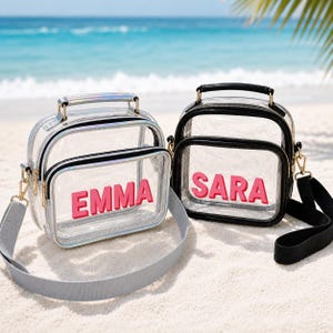 May include: Two clear crossbody bags with top handles and adjustable straps. The left bag has silver trim and the name EMMA in pink letters. The right bag has black trim and the name SARA in pink letters. Both bags feature a front zippered pocket.