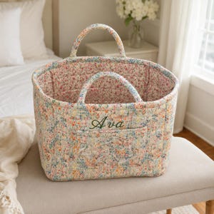May include: A quilted fabric basket featuring a colorful floral pattern with small pink, blue, and yellow flowers. The basket has two rounded handles and a front pocket embroidered with the name Ava in dark green cursive script lettering.