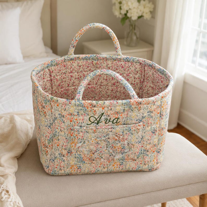 May include: A quilted fabric basket featuring a colorful floral pattern with small pink, blue, and yellow flowers. The basket has two rounded handles and a front pocket embroidered with the name Ava in dark green cursive script lettering.