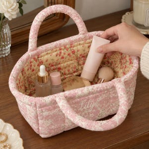 May include: A quilted fabric storage basket featuring a pink and white floral pattern with two matching handles. The exterior is embroidered with the name Lily in white script. The interior is lined with a cream fabric featuring small red flowers.