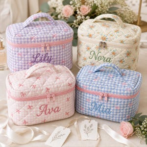 Peut inclure: Quatre trousses de toilette rectangulaires matelassées avec poignées supérieures et doubles fermetures éclair. Une est à carreaux violets avec le nom Lily, une est crème à motifs floraux avec Nora, une rose à motifs nœuds avec Ava, et une à carreaux bleus avec Mia.