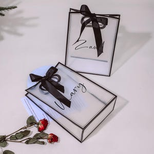 Puede incluir: Dos bolsas de regalo de pl&aacute;stico transparente con lazos de cinta negra y letras negras. Las bolsas est&aacute;n atadas con una cinta negra y tienen los nombres "Zavi" y "Harry" escritos en ellas.