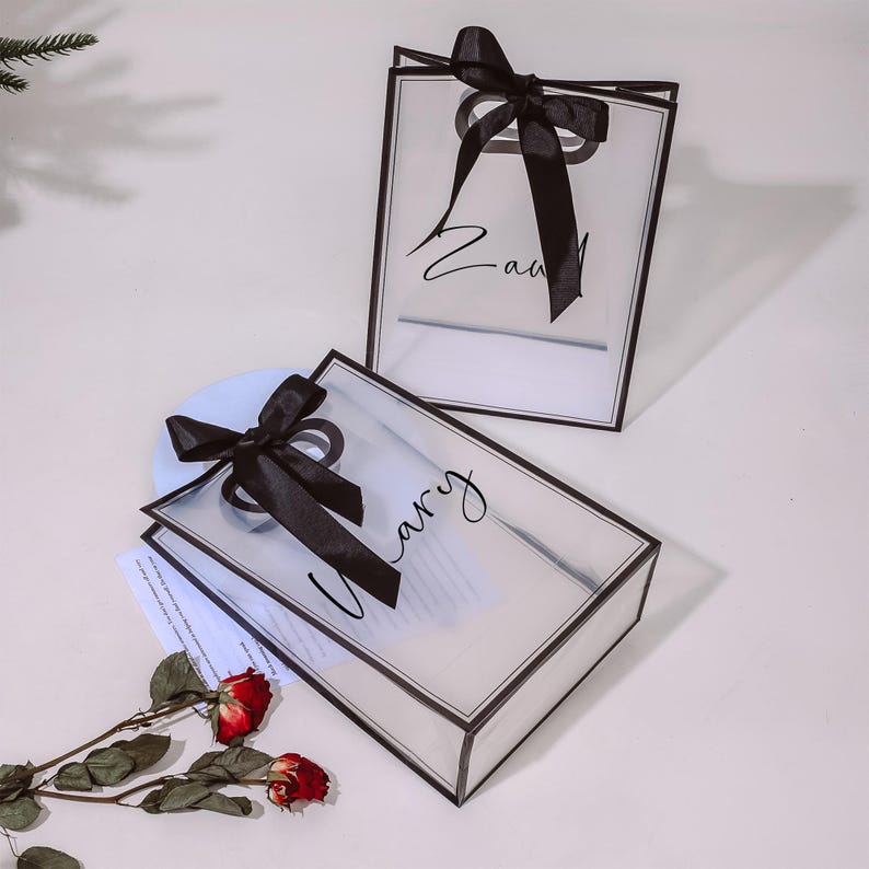 Puede incluir: Dos bolsas de regalo de pl&aacute;stico transparente con lazos de cinta negra y letras negras. Las bolsas est&aacute;n atadas con una cinta negra y tienen los nombres "Zavi" y "Harry" escritos en ellas.
